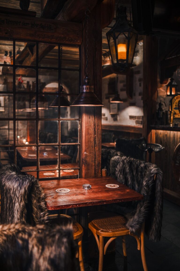 Przytulne wnętrze Frost Bar w hotelu Paul’s Fantasia Boutique Suites w Szczecinie. Rustykalny wystrój z drewnianymi belkami, kamiennymi ścianami i nastrojowym, ciepłym oświetleniem. W lokalu stoją drewniane stoły i krzesła pokryte ciemnymi futrzanymi narzutami, a przy oknach wiszą industrialne lampy, tworząc kameralną atmosferę.