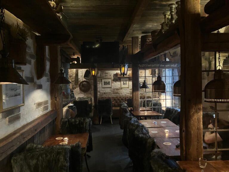 Przytulne wnętrze Frost Bar w hotelu Paul’s Fantasia Boutique Suites w Szczecinie. Rustykalny wystrój z drewnianymi belkami, kamiennymi ścianami i nastrojowym, ciepłym oświetleniem. W lokalu stoją drewniane stoły i krzesła pokryte ciemnymi futrzanymi narzutami, a przy oknach wiszą industrialne lampy, tworząc kameralną atmosferę.